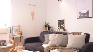 A cozy living room with a dark grey sofa, a small wooden coffee table with a book and croissants, a side table with a guitar and a framed picture, and a wall with a cat illustration and a chalkboard sign.