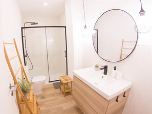 A modern bathroom with a glass shower enclosure, a wooden ladder, a small stool, and a round mirror above a white sink with a wooden cabinet.