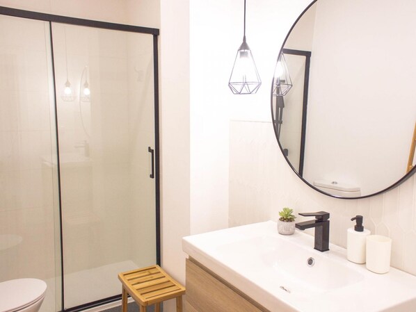 A modern bathroom with a glass shower enclosure, a wooden stool, a white sink with a black faucet, a round mirror, and pendant lighting.