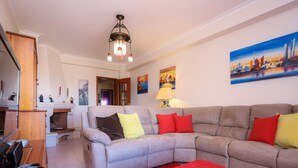 A living room with a grey sofa, red and yellow cushions, a red rug, a chandelier, and a cityscape painting on the wall. - Brasil Avenue by Rent4all (Figueira da Foz)