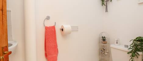 Un baño con inodoro, un rollo de papel higiénico, una toalla rosa colgada en un perchero y un pequeño estante con una planta y un rollo de papel higiénico.