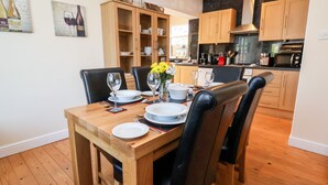 Un coin repas avec une table en bois dressée pour quatre personnes, entourée de chaises noires. On aperçoit, au fond, un buffet en bois avec des portes en verre, ainsi qu’un espace cuisine avec des meubles et des appareils électroménagers en bois.