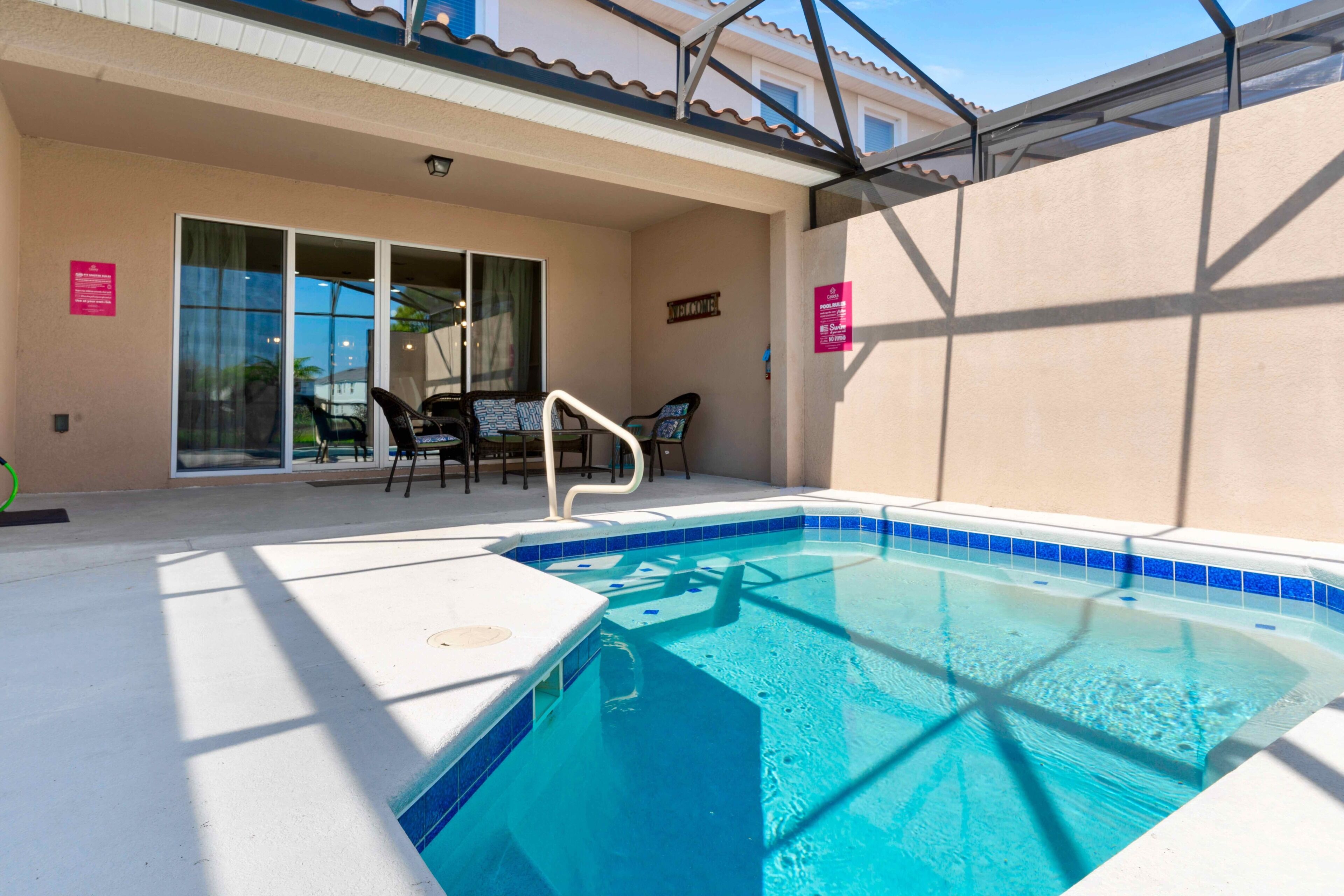 Ein Swimmingpool mit Fliesenrand, eine überdachte Terrasse mit Stühlen und eine Glasgleittür zu einem Außenbereich.