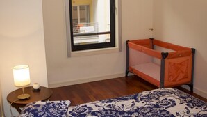 Una habitación con ventana, una cama con colcha estampada, una mesita de noche con lámpara y una cuna con mosquitero naranja.