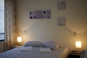 Une chambre à coucher avec une couvre-lit à motifs floraux, deux lampes de chevet, une œuvre d’art accrochée au mur et une fenêtre ornée de rideaux à fleurs.