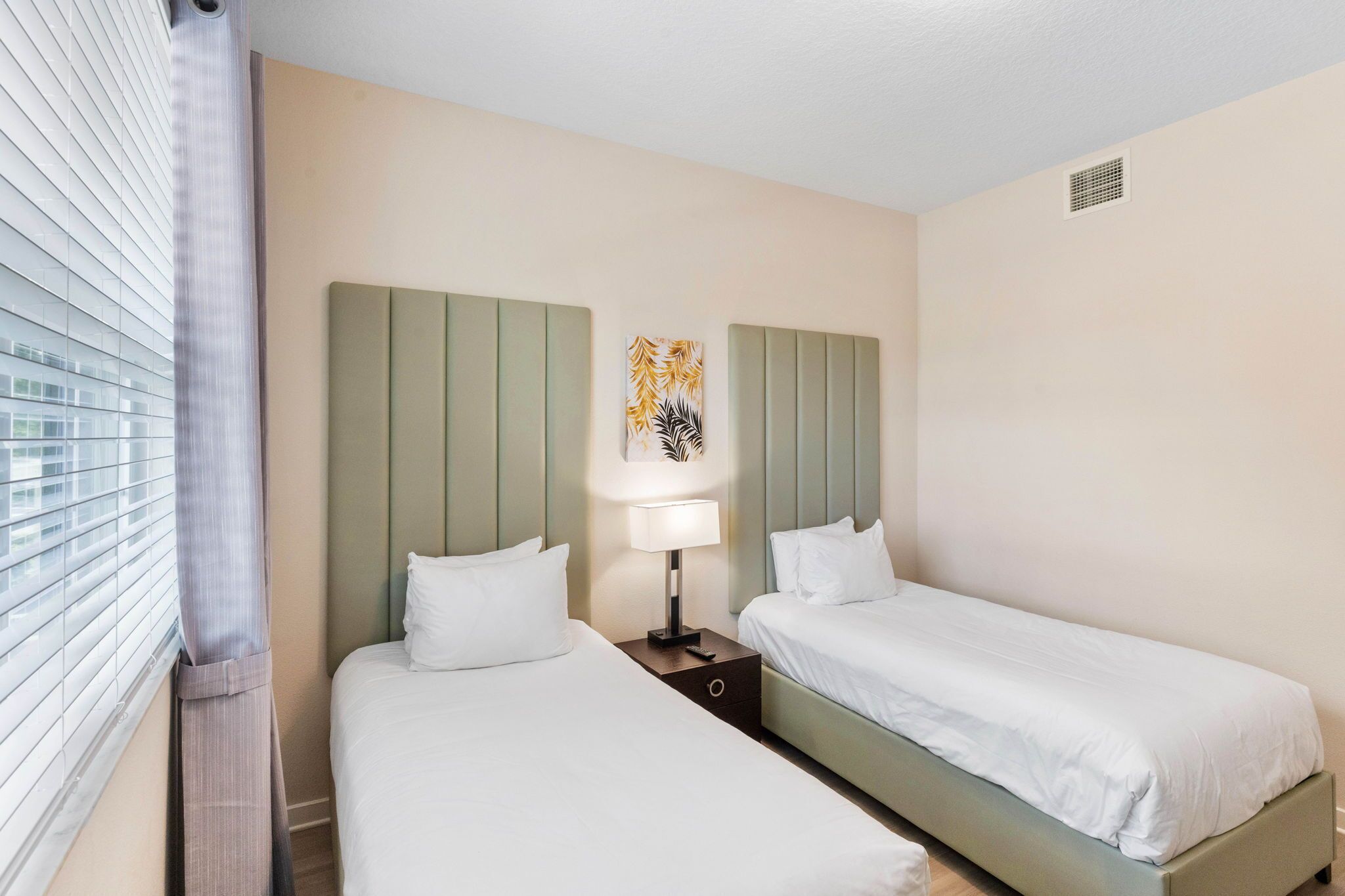 A hotel room with two beds, a nightstand with a lamp, and a window with blinds.