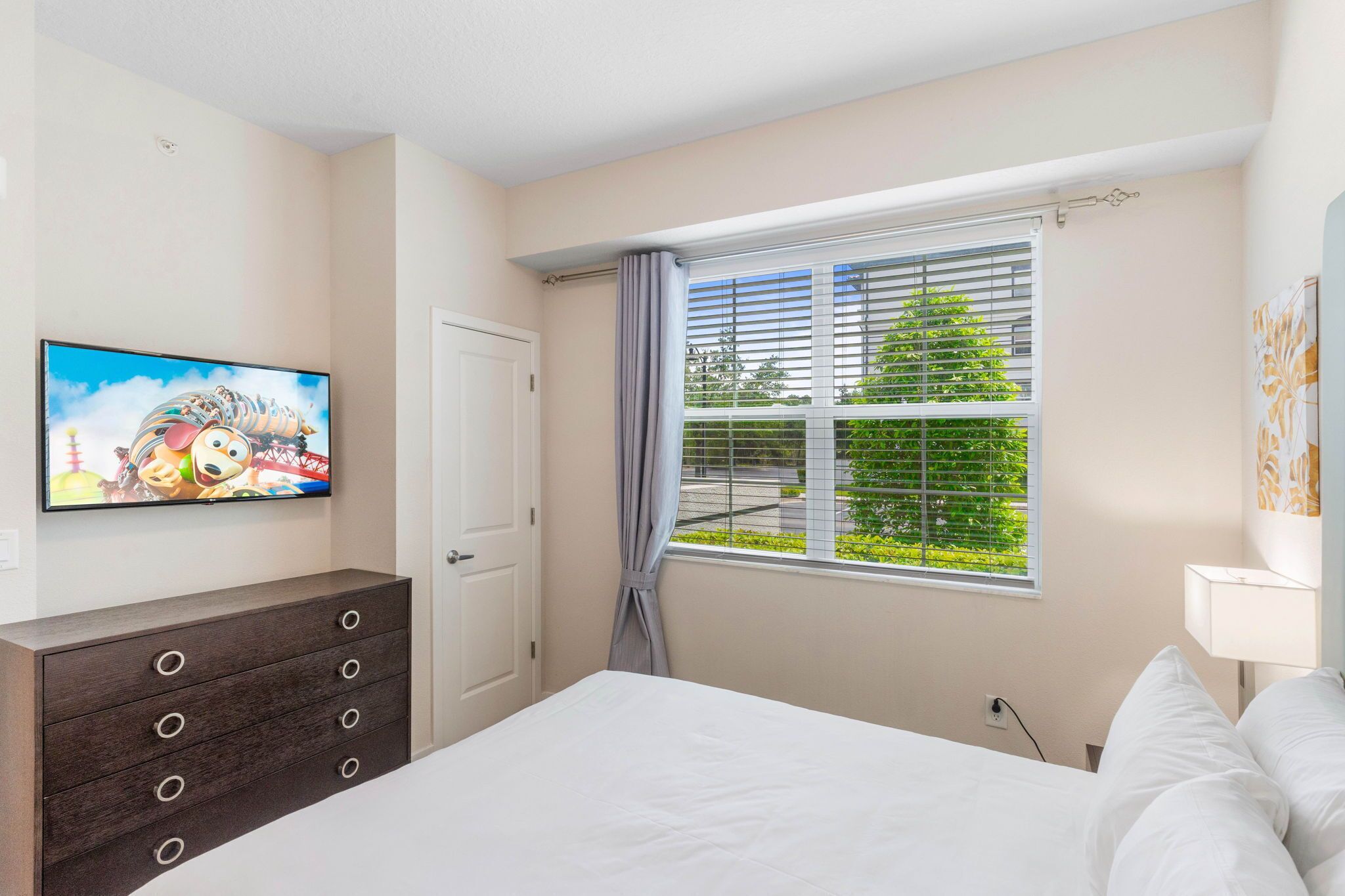 A bedroom with a bed, a TV on the wall, a window with blinds, and a dresser.