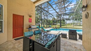 Um pátio coberto com estrutura de vidro, mobiliário externo, churrasqueira e piscina.