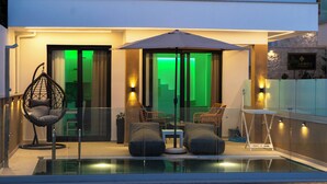 A modern outdoor pool area with a glass enclosure, cushioned seating, and ambient lighting. - Vrachos Luxury Maisonettes (Parga)
