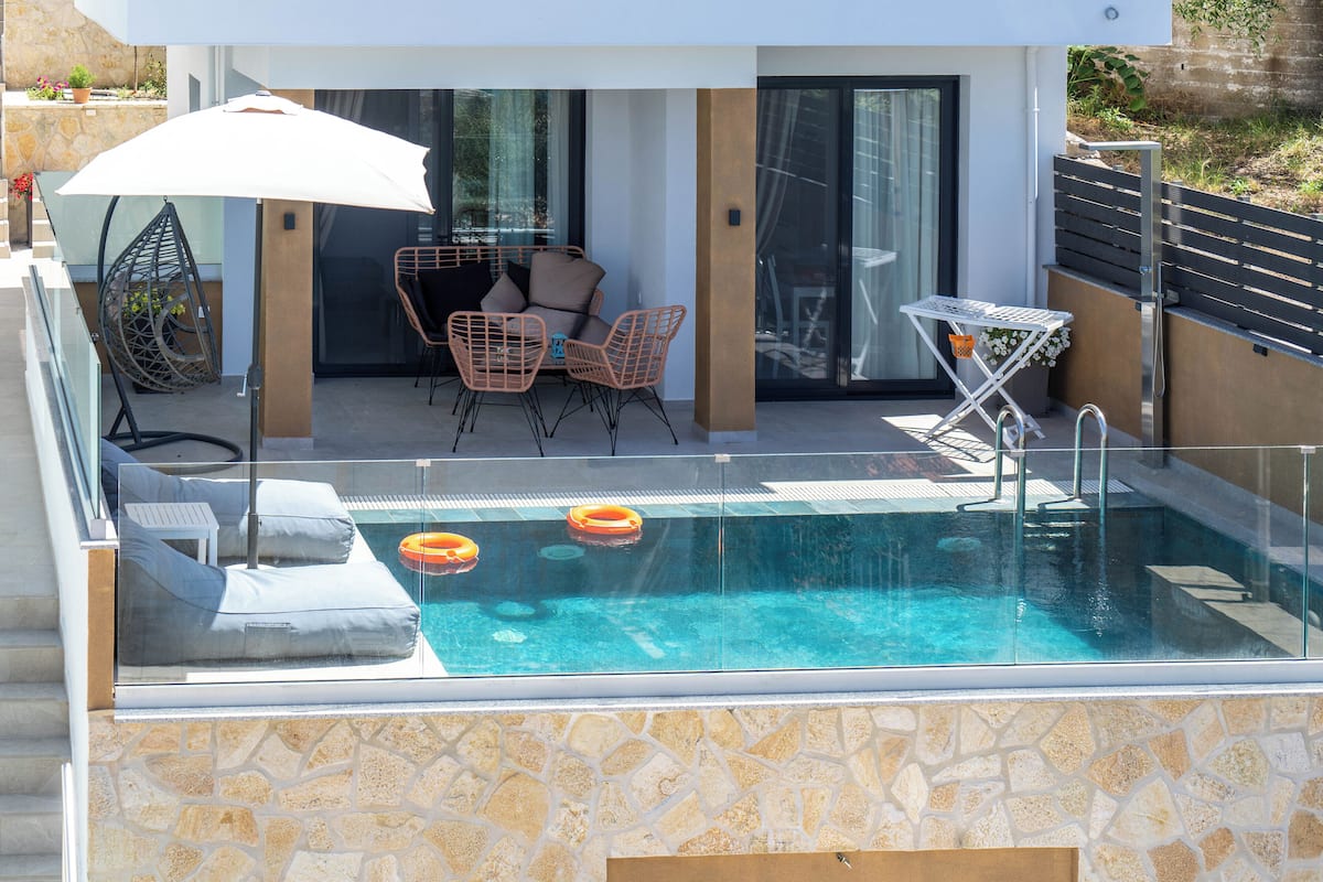A modern outdoor pool area with a glass balustrade, lounge chairs, and a table with an umbrella.