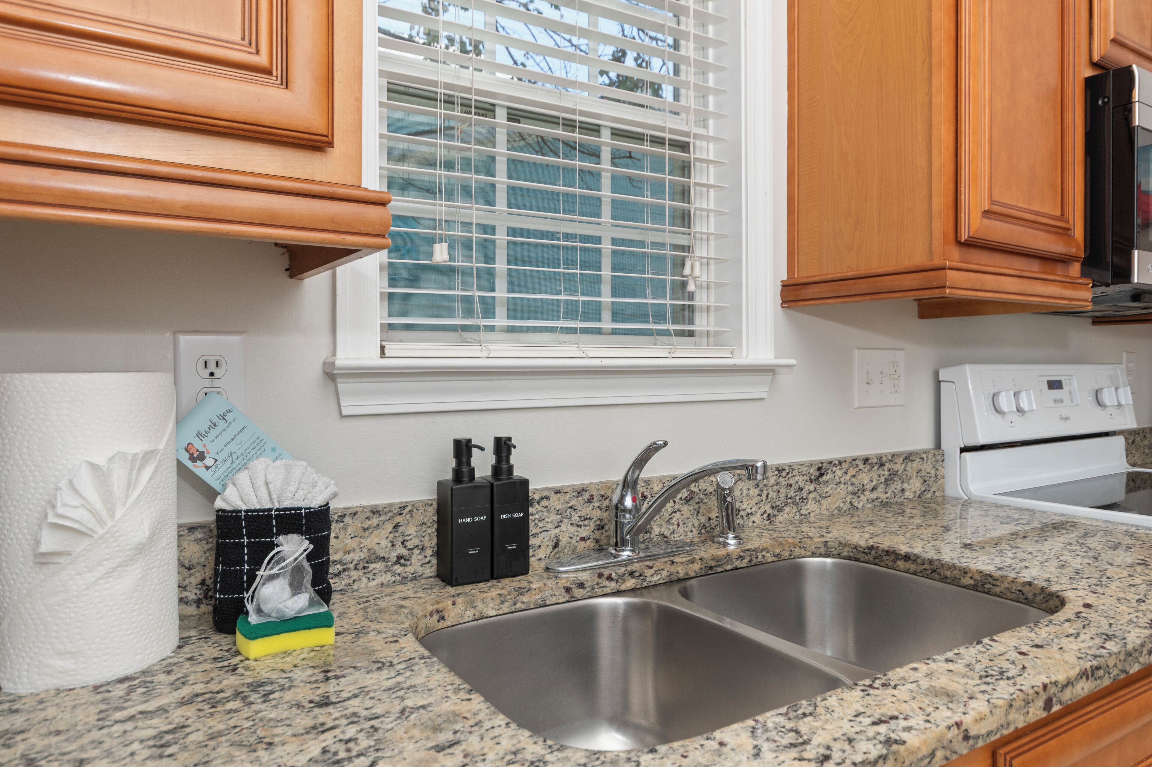 A kitchen with granite countertops, a stainless steel sink, wooden cabinets, a window with blinds, and a white stove.