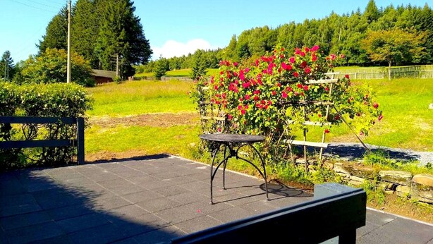 An outdoor seating area with a bench and a small table, surrounded by a garden with blooming flowers and a view of a green field and trees. - Ferienhaus in Barenstein bei Grunen Hugeln (Baerenstein)