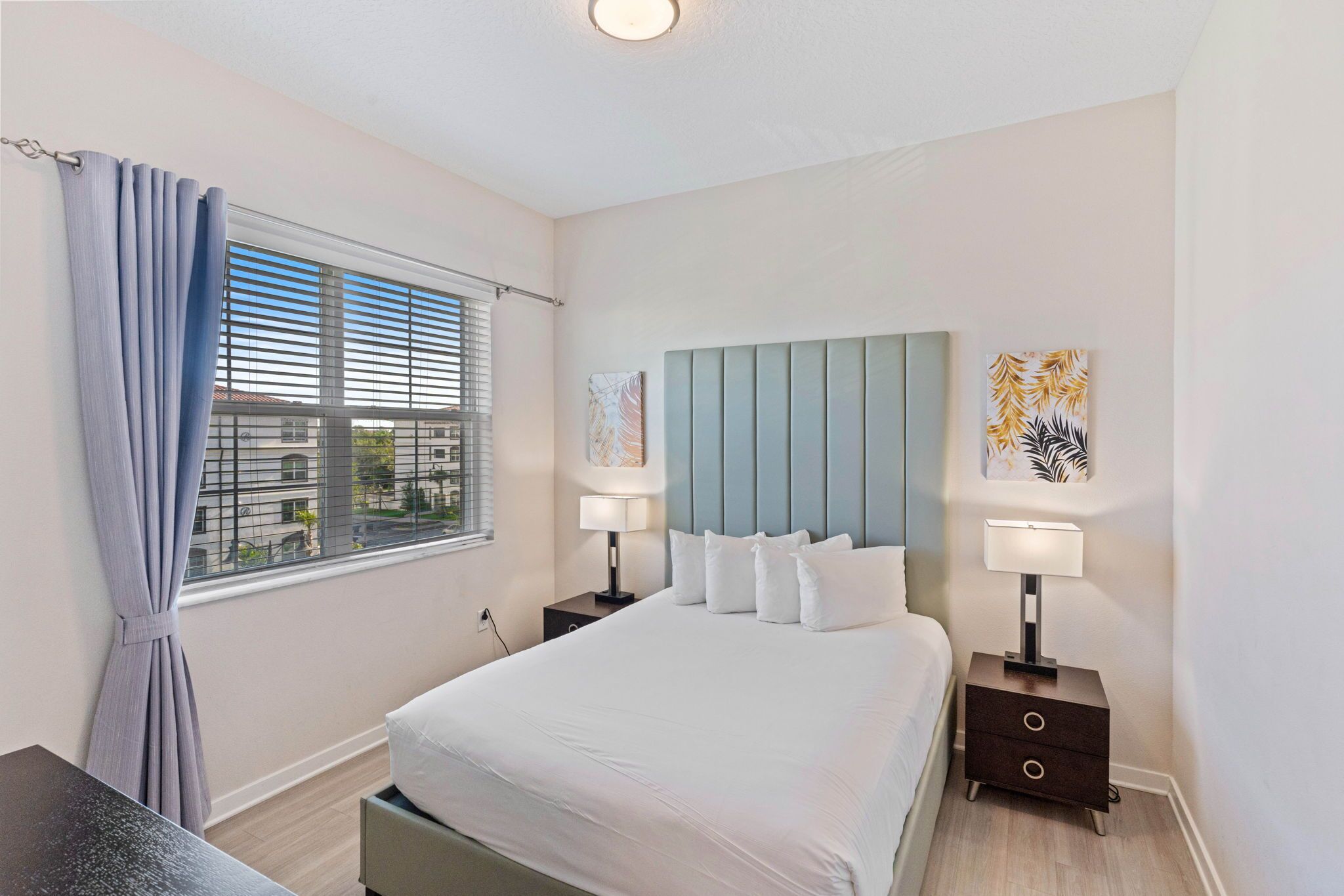 A bedroom with a large bed, two bedside tables with lamps, a window with blinds, and artwork on the wall.