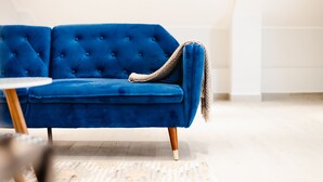 Un canapé bleu en velours avec des pieds en bois, un plaid tissé et une petite table ronde à la surface blanche.