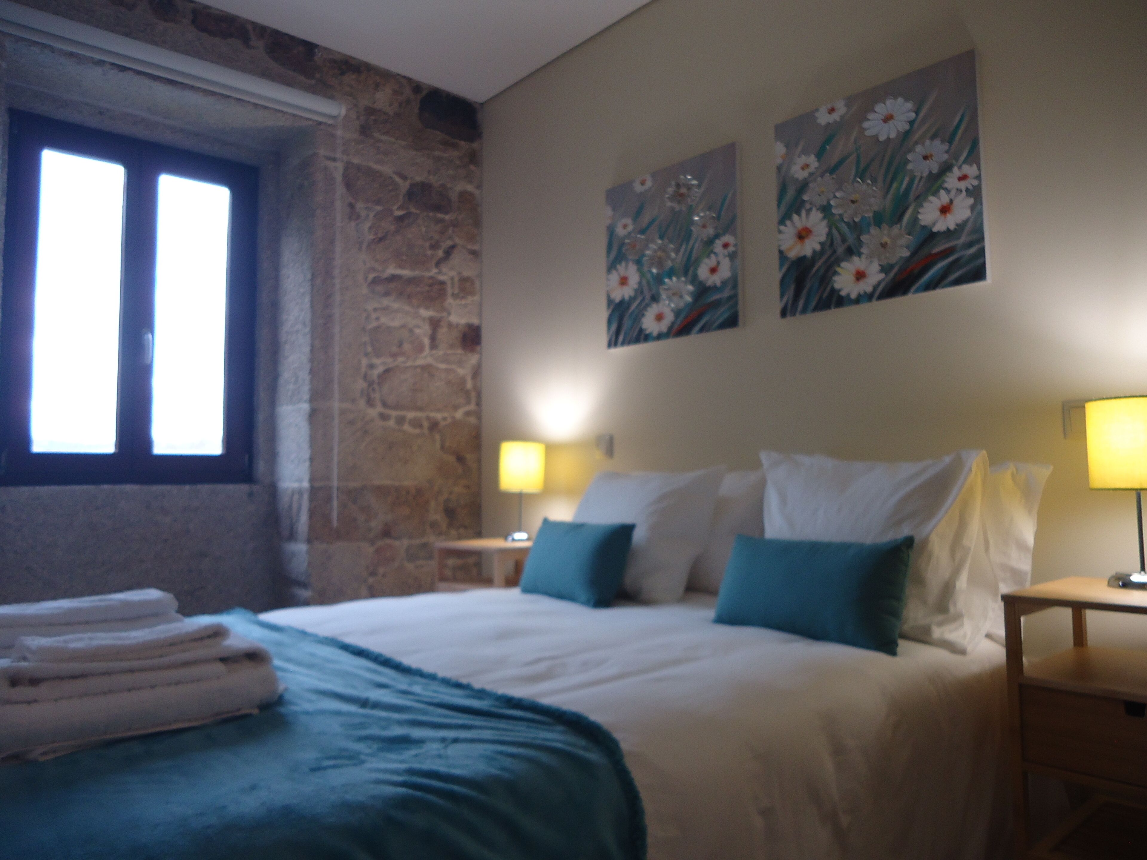 Une chambre avec un mur en pierre, un lit avec une literie blanche et bleue, deux lampes fixées au mur et une fenêtre avec des stores.