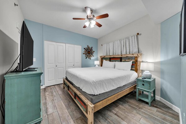 A bedroom with a wooden bed, white bedding, a ceiling fan, a TV on a cabinet, and a small bedside table.