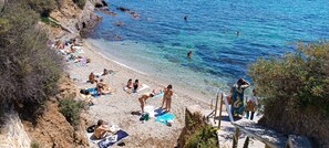 Ein Strand mit badenden Personen, klarem blauen Wasser und felsigem Gelände.