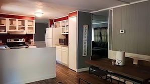 A compact kitchen with white cabinets, a stove, and a microwave. There's a dining area with a table and benches. A sign on the wall reads "LAKE HOUSE." - Near The Lake (Bee Branch)