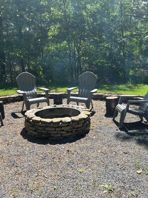 Relax in a serene outdoor setting with a stone fire pit surrounded by Adirondack chairs amidst lush greenery.