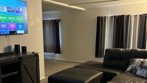 A living room with a large flat-screen TV mounted on the wall, a black leather sofa, a white coffee table, and a dark leather ottoman. - Near The Lake (Bee Branch)