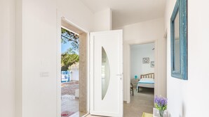 Un couloir mène à une chambre à coucher qui contient un lit, une table de chevet ornée d’un vase de fleurs et une porte donnant sur l’extérieur.
