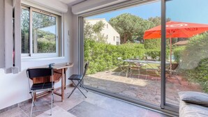 Une pièce dotée d’une porte-fenêtre coulissante donnant sur un patio extérieur avec une table et des chaises, un parasol rouge, et une vue sur la verdure et un bâtiment.