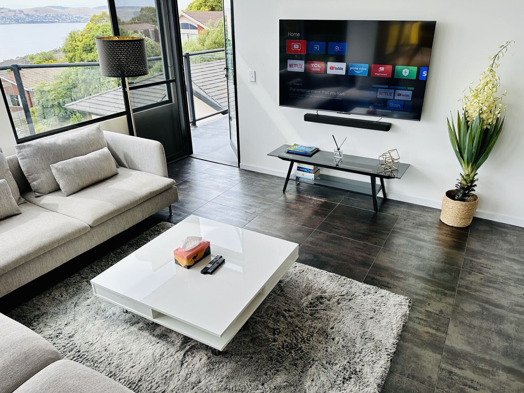 A modern living room with a large TV mounted on the wall, a sofa, a coffee table, and a view of the outdoors.