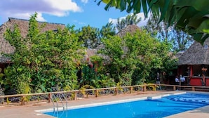 Une piscine dotée d’une terrasse en bois, entourée d’une végétation luxuriante et bordée de constructions aux toits de chaume.