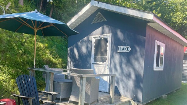 Uma pequena cabana com uma placa 'LAKE', um deck de madeira, cadeiras de vime e um guarda-sol azul.