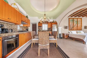 A kitchen with wooden cabinets, a dining table with wicker chairs, a chandelier, and a bed with a view of palm trees.