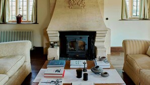 A cozy living room with a stone fireplace, two windows with curtains, a radiator, and a coffee table with books and decorative items.