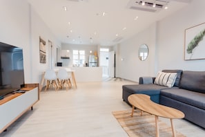 A modern living room with a grey sofa, a wooden coffee table, and a TV on a wooden stand. There is a dining area with a white table and chairs in the background.