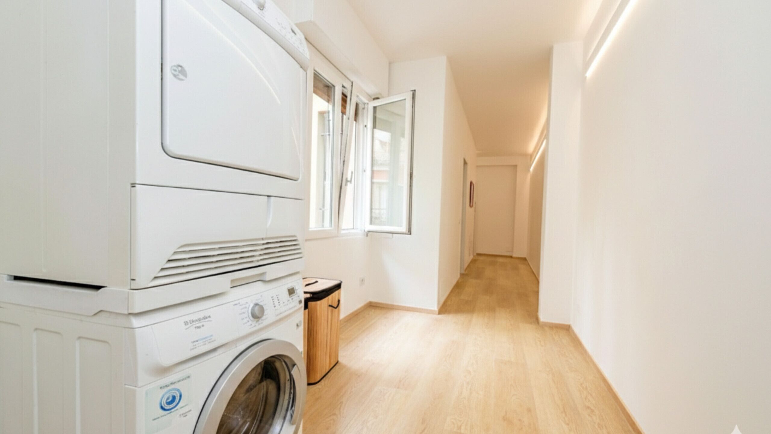 A laundry area with a front-loading washing machine and dryer, a wooden bench, and a window with curtains.