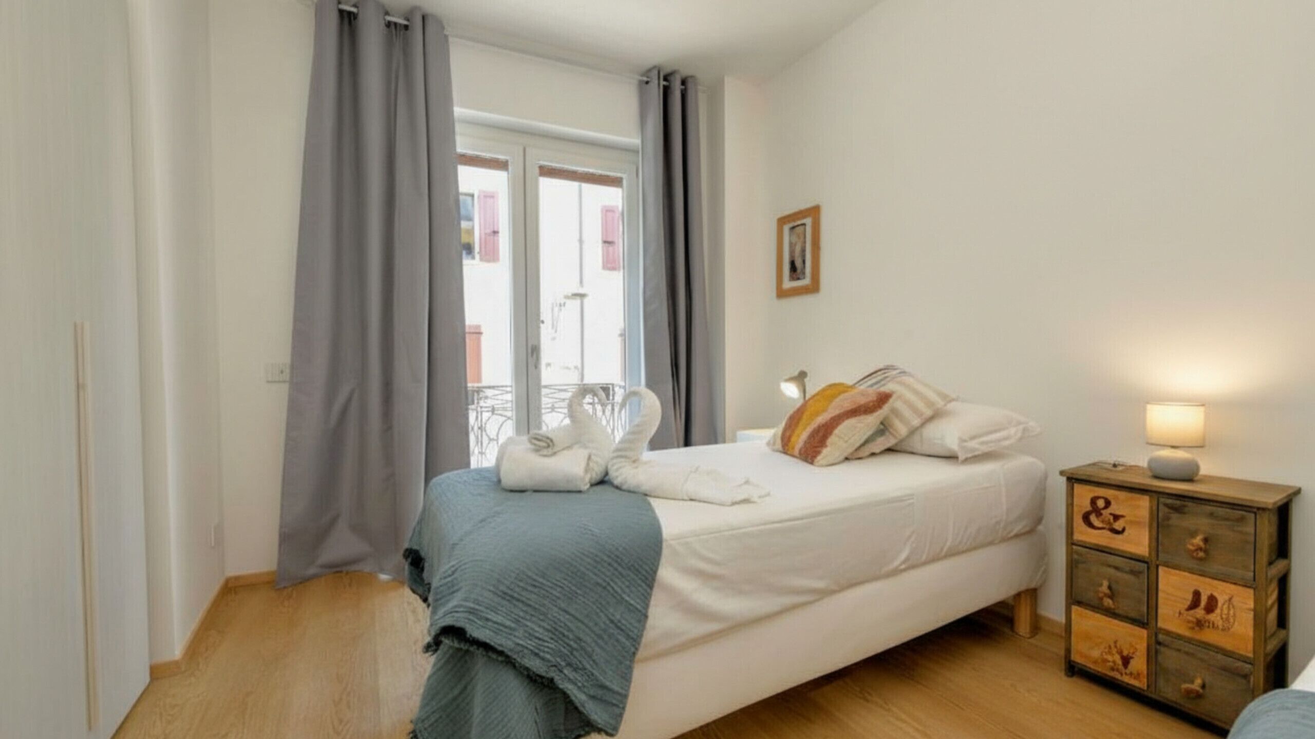 Ein Schlafzimmer mit einem Bett, einem Nachttisch mit Lampe, einer Kommode und einem Fenster mit Vorhängen.