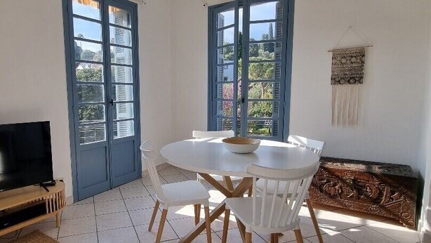 A dining area with a round table and chairs, a television on a wooden stand, and large windows with blue shutters. - Bright Appartment, perfect location (Saint-Jean-Cap-Ferrat)