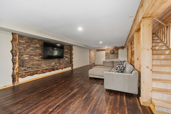 A spacious living room with a stone accent wall, a flat-screen TV mounted on it, and a wooden staircase on the right.