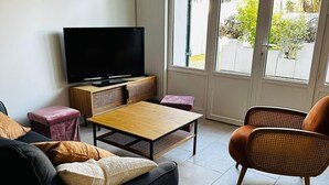 A modern living room with a flat-screen TV, a sofa, a coffee table, and a wicker armchair.