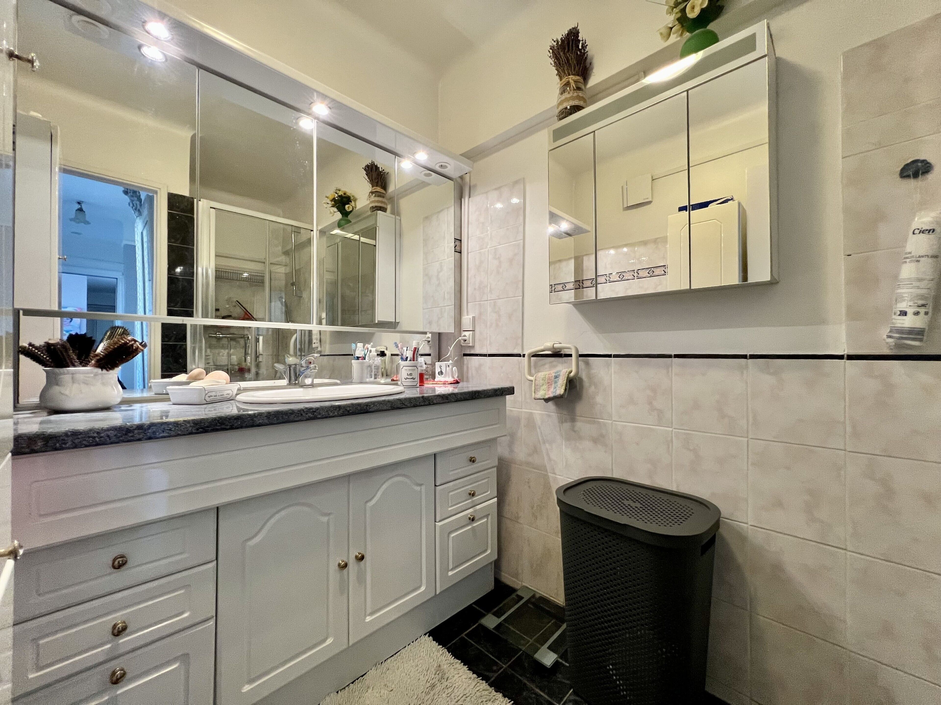 Une salle de bain moderne avec un grand miroir, des meubles blancs et une poubelle noire.