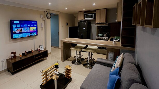 Een moderne woonkamer met een flatscreen-tv, een bank, een salontafel en een keukenhoek met een magnetron en een koelkast.