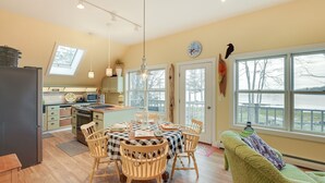 A cozy kitchen with a dining area, wooden chairs, a table with a checkered tablecloth, and a view of the outdoors through large windows. - Waterfront Deer Isle Apartment w/ Fire Pit (Deer Isle)