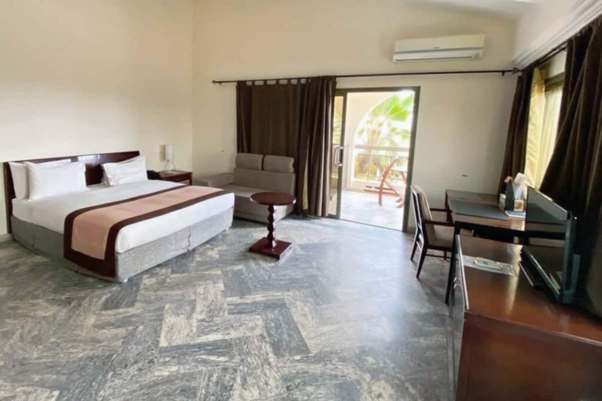 A hotel room with a bed, a sofa, a small table, a desk, and a chair. There is a window with curtains, and an air conditioning unit on the wall.