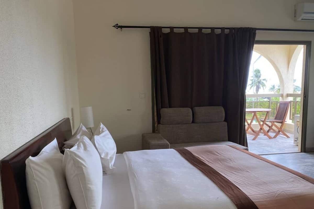 A hotel room with a large bed, a sofa, a balcony with outdoor furniture, and a view of palm trees.