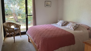 A bedroom with a bed, a wicker chair, and a view of greenery outside. - La Girolle (Lège-Cap-Ferret)