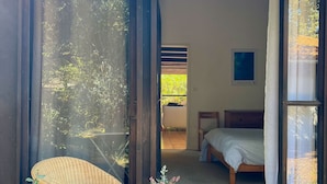 A wooden deck with a wicker chair and a small table, a sliding glass door leading to a bedroom with a bed and a nightstand, and a framed picture on the wall. - La Girolle (Lège-Cap-Ferret)