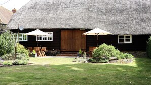 Une petite maison au toit de chaume, avec une porte en bois, deux fenêtres à cadre blanc et un petit porche équipé d’une table et de chaises.