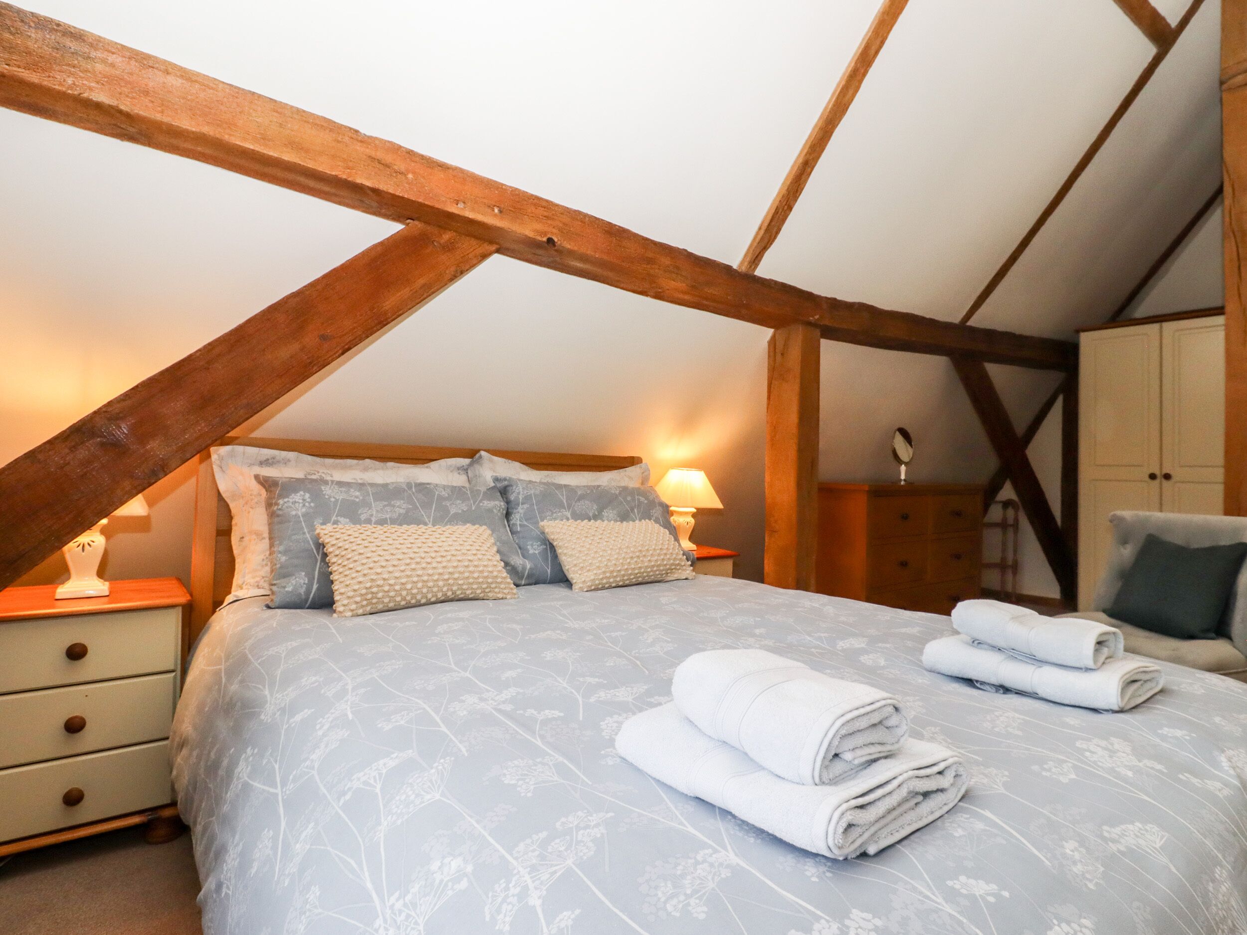 A bedroom with a sloped ceiling, wooden beams, a bed with floral bedding, two bedside tables with lamps, a dresser, and a chair.