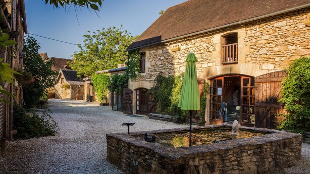 A courtyard with a stone fountain, a green umbrella, and stone buildings with arched windows. - Charming cottage within a perigordine farmhouse  (Saint-Cyprien)