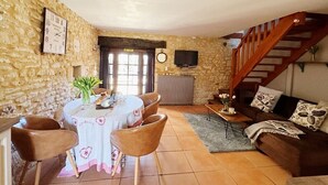 A cozy living room with a stone wall, a dining table with chairs, a sofa, a TV, and a staircase. - Charming cottage within a perigordine farmhouse  (Saint-Cyprien)