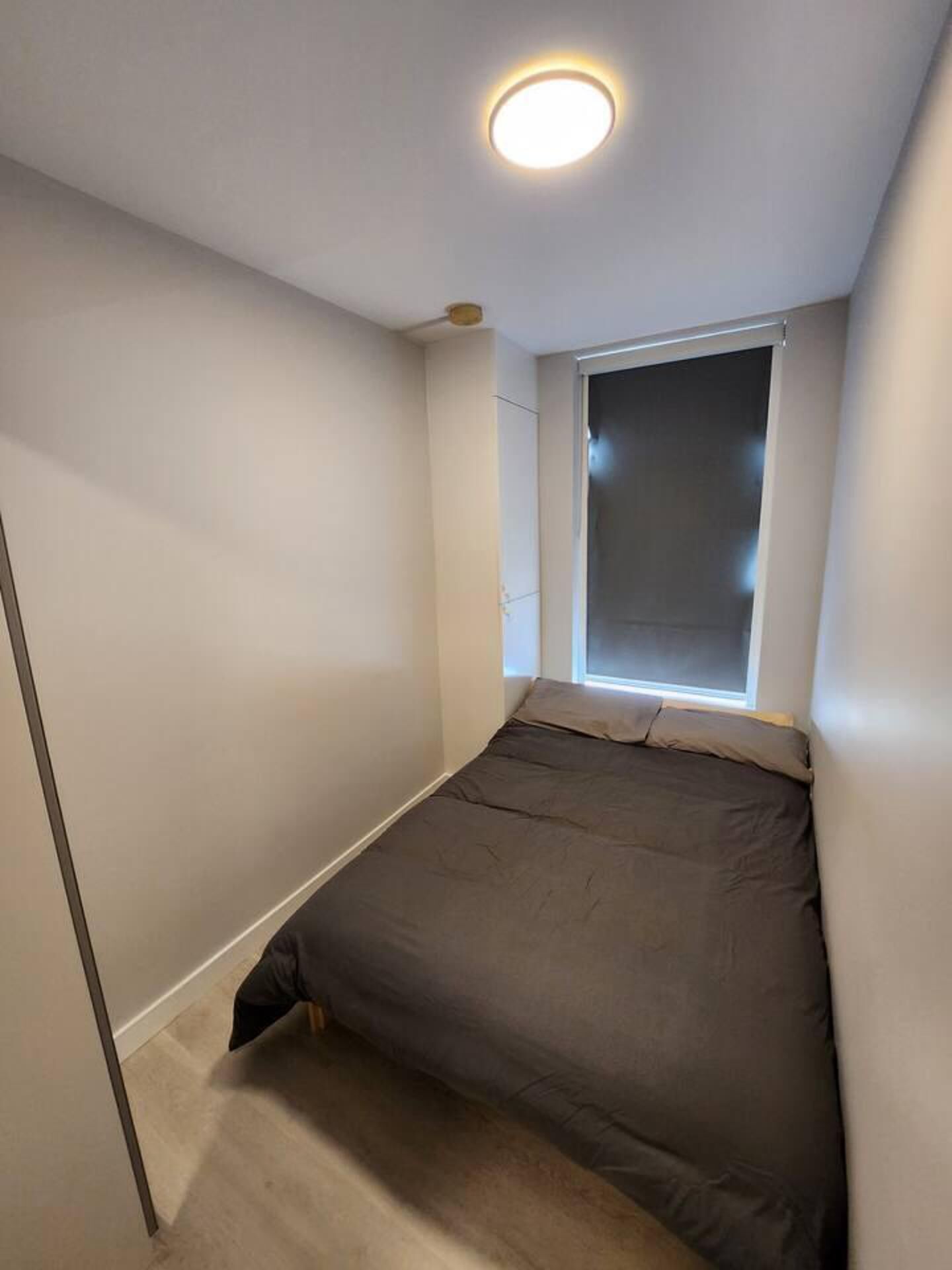 A small, narrow room with a single bed, a window, and a ceiling light.