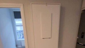 A hallway with a wooden floor, a white wall-mounted electrical box, and a door leading to another room. - Beautiful 2-bed Apartment in London - Sleeps 6! (London)